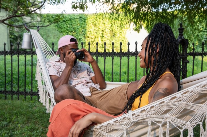 A couple relaxes in a hammock in a backyard setting. One African-American man is taking a photo with a DSLR camera, focusing on the woman.