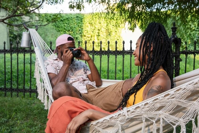 A couple relaxes in a hammock in a backyard setting. One African-American man is taking a photo with a DSLR camera, focusing on the woman.
