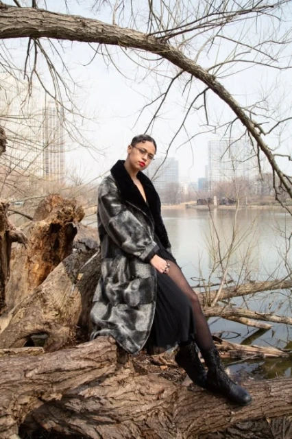 Woman in fur coat posing on tree trunk near lake in urban park setting with city buildings in background, fashion editorial style.