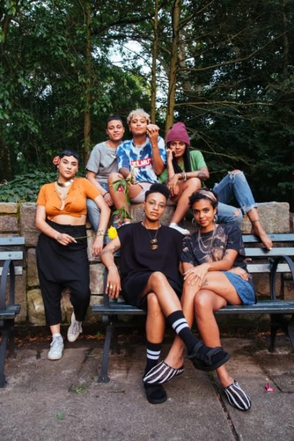 Six diverse young adults pose on a park bench surrounded by lush green trees, exuding a relaxed and natural vibe in a group portrait.