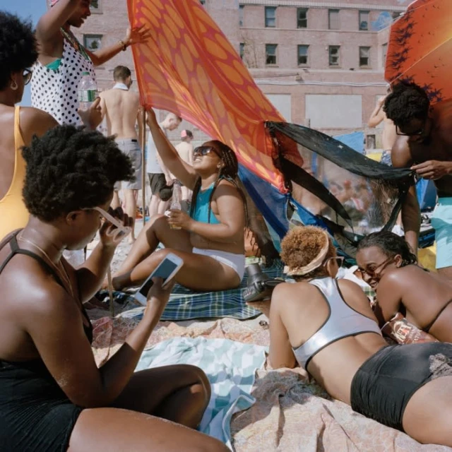 Diverse group of people relaxing on a sandy beach with a brick building in the background, some holding up an orange fabric.