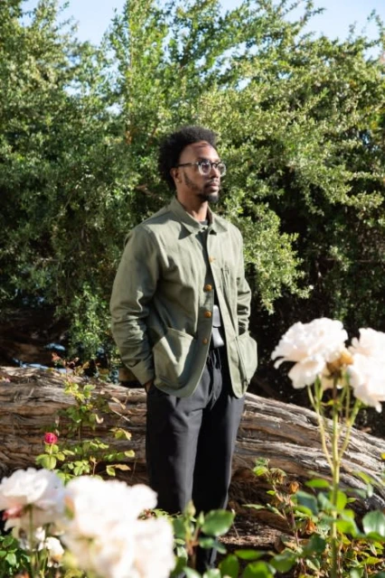 Adult male with an afro and glasses stands outside wearing an olive green chore coat, white shirt, and black pants in a sunny garden.