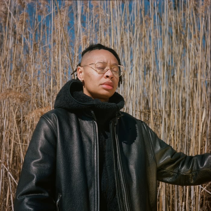 Portrait of a non-binary person with short dreadlocks and glasses in a black jacket against a backdrop of tall reeds under a blue sky.