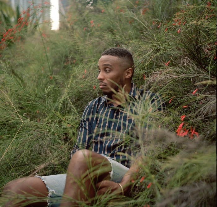 Black man seated amidst tall green grass wearing a striped shirt and light shorts