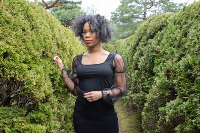 An African-American woman poses in a black dress among green hedges in a garden setting, captured in an eye-level, medium shot.