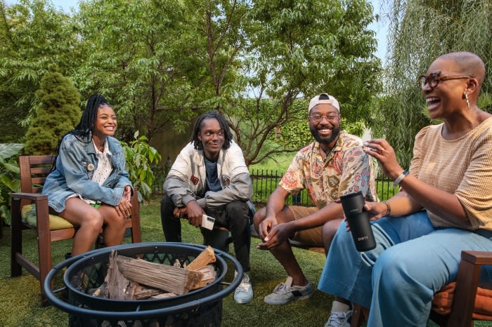 Group of four young adult and elderly adults sitting in wooden chairs outdoors on a lawn, chatting and smiling near a fire pit.