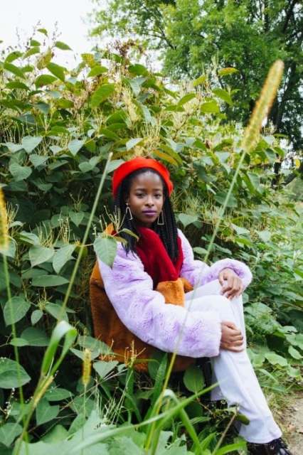 A young woman sits surrounded by lush green plants, wearing a colorful fur jacket and a red beret, looking thoughtfully at the camera.