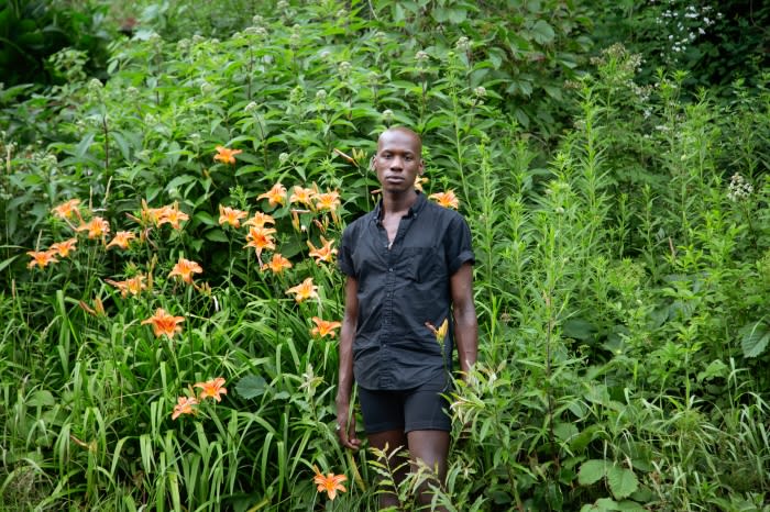Adult male standing in front of orange lilies and green foliage, wearing a short-sleeved black shirt and black shorts in an outdoor setting.