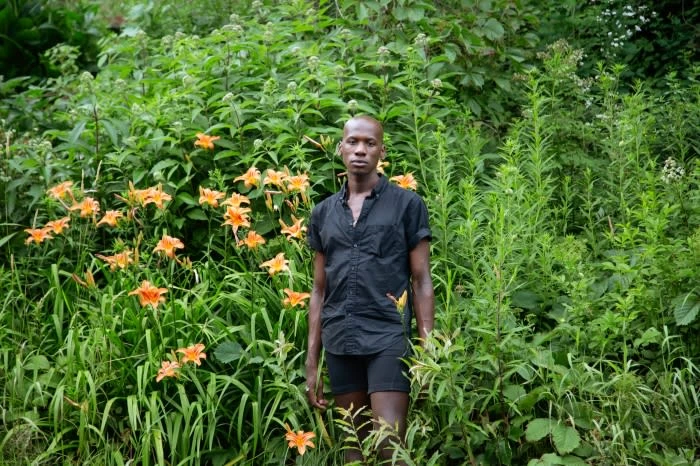 Adult male standing in front of orange lilies and green foliage, wearing a short-sleeved black shirt and black shorts in an outdoor setting.