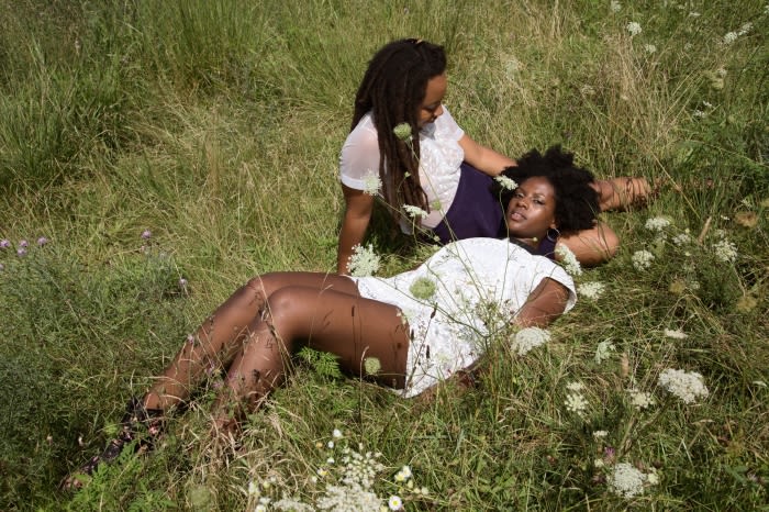 Two dark-skinned women pose together in a grassy field on a sunny day. One woman lies down while the other sits and touches her shoulder.