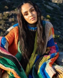 Portrait of a woman with long dark hair wearing a vibrant multicolored crochet sweater outdoors against a natural mountain hillside backdrop.