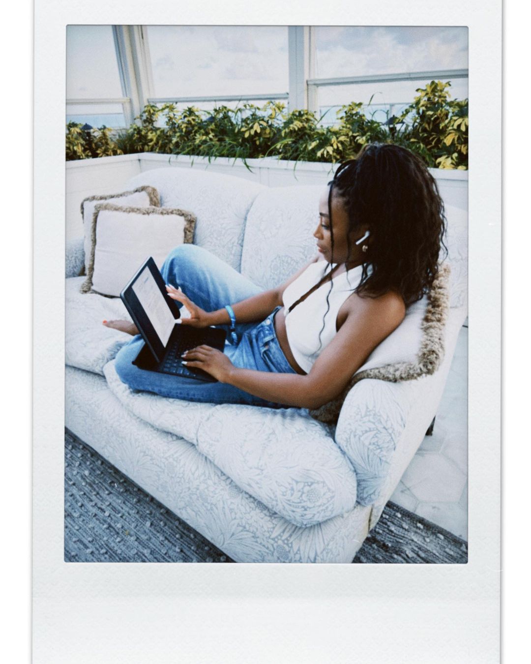 Black woman seated on a patterned sofa using a tablet with a keyboard in a bright room by Naima Green