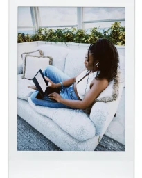 Black woman seated on a patterned sofa using a tablet with a keyboard in a bright room by Naima Green