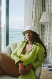 Black woman wearing a white bucket hat and lime green jacket sitting in an upholstered chair near a window with an ocean view.