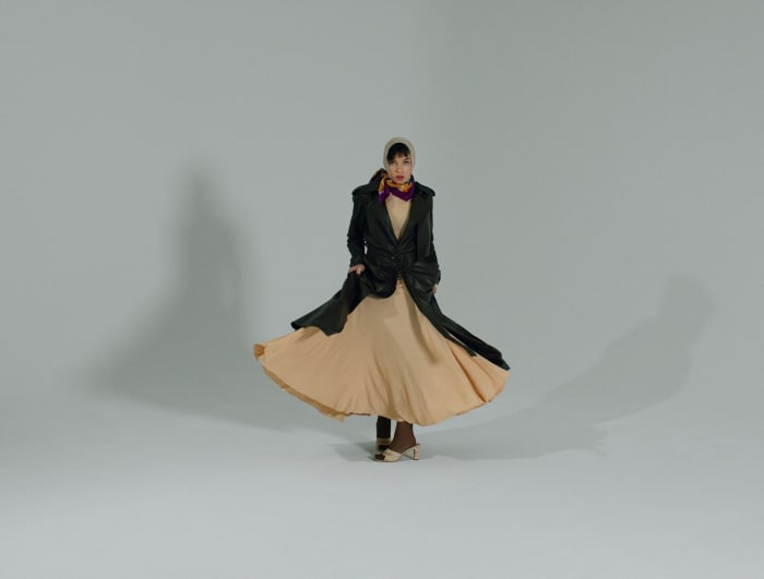 Young woman in black coat and beige dress twirling in photo studio. She wears a cream headscarf and brown heeled shoes against grey background.