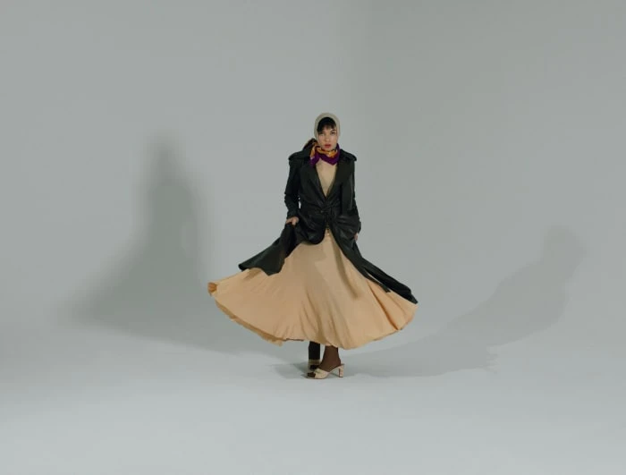 Young woman in black coat and beige dress twirling in photo studio. She wears a cream headscarf and brown heeled shoes against grey background.