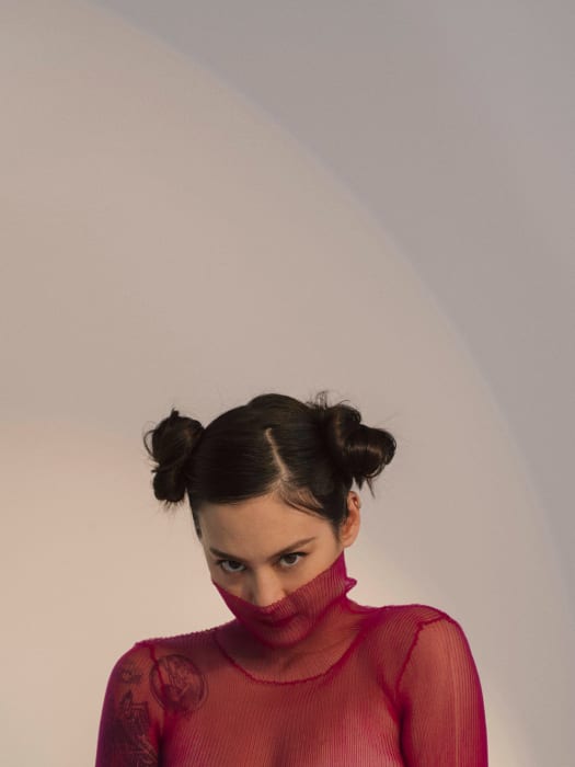 A young adult woman with dark hair styled in space buns, wearing a red mesh top, gazes intensely at the camera against a neutral backdrop.
