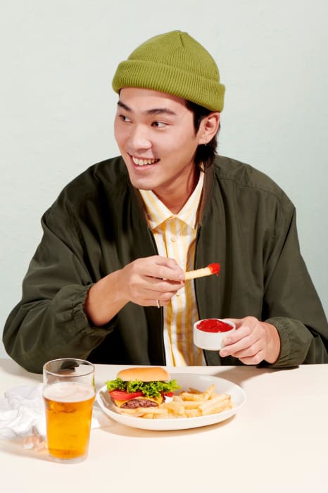 Young man in olive beanie smiles, dips french fry in ketchup, with burger and beer on table in front of pale green backdrop.