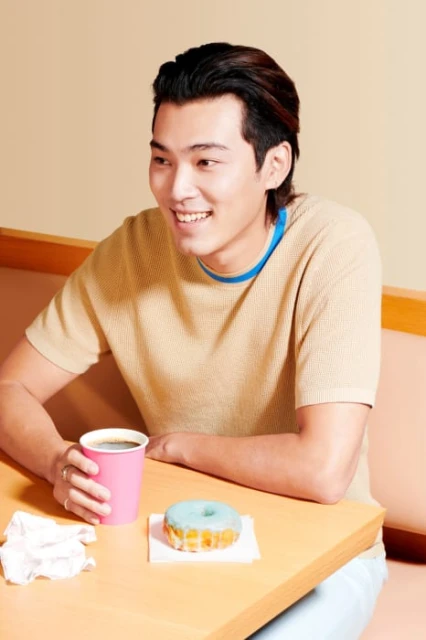 Young Asian man smiles at a table with a coffee cup and teal-iced donut. Indoor shot with natural lighting, light peach background.