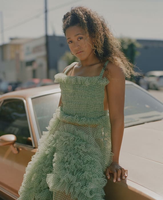 A young woman with curly hair in a green dress leans against a tan car in an urban setting on a sunny day. Lifestyle fashion.
