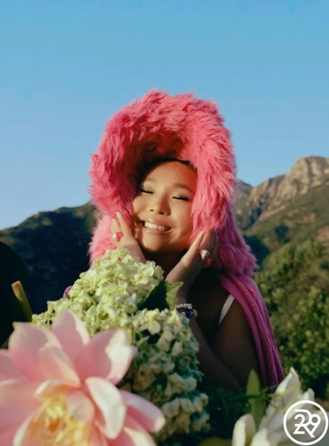 Woman with pink faux fur hood and flower bouquet in mountain landscape under blue sky. Smiling woman with hands near face.