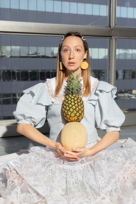 Woman in vintage-style dress sits indoors in front of a window, balancing a pineapple on top of a cantaloupe melon in her hands.