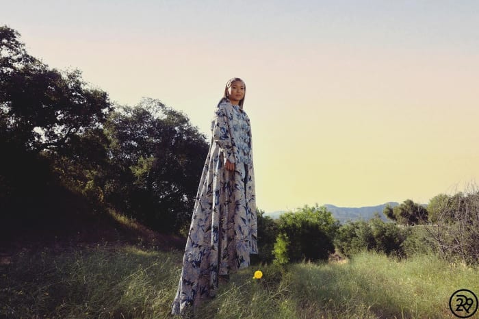 Woman in long, floral dress stands in a grassy field with trees under a sunny sky, fashion in nature.