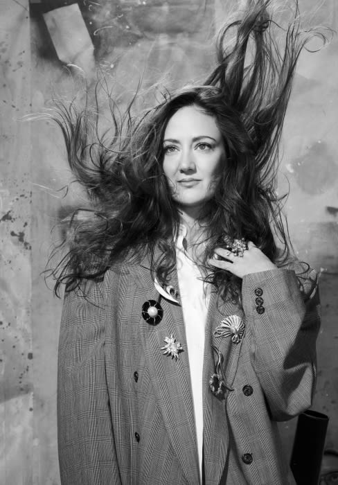 Black and white portrait of a woman in a blazer, with hair thrown upward, indoor with mottled background and soft lighting. Fashion editorial.