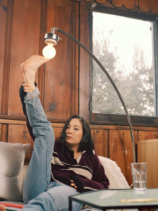 Woman sits by a lamp, her foot touching the bulb, with a lighthearted expression. The room has wood paneling and a window.