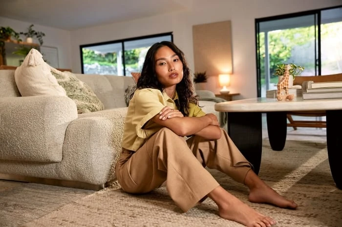 A young woman sits barefoot on a rug in a light-filled living room with a tan couch and large sliding glass doors to the outside.
