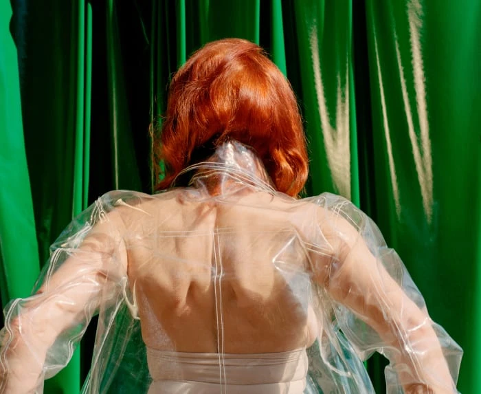 Back view of a redhead woman in a translucent jacket posing against a vibrant green backdrop, showcasing fashion and style.