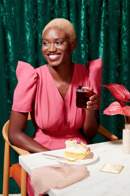 Smiling Black woman in a pink dress enjoys a drink at a white marble table with cake, set against a dark green crushed velvet backdrop.
