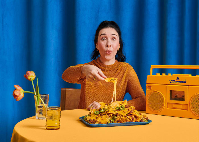 Adult female eats cheesy nachos at a table covered with an orange tablecloth, in front of a blue curtain backdrop, with a yellow radio.