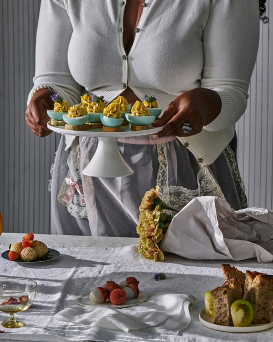Woman serves deviled eggs on cake stand surrounded by fruit and flowers on linen tablecloth in a neutral light indoor setting.