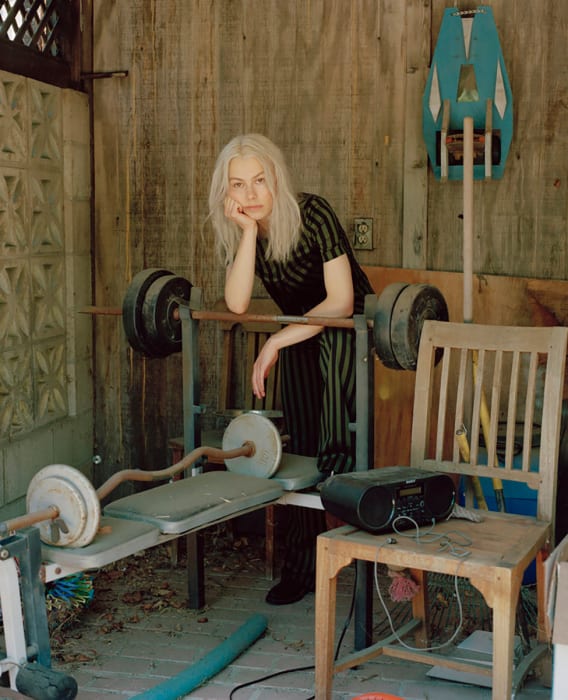 Phoebe Bridgers in black and green striped dress leans against weight bench in cluttered, weathered indoor setting with music equipment.