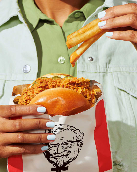 A person holds a chicken sandwich and french fries in a KFC paper bag. The person is wearing a green jacket and light blue nail polish.
