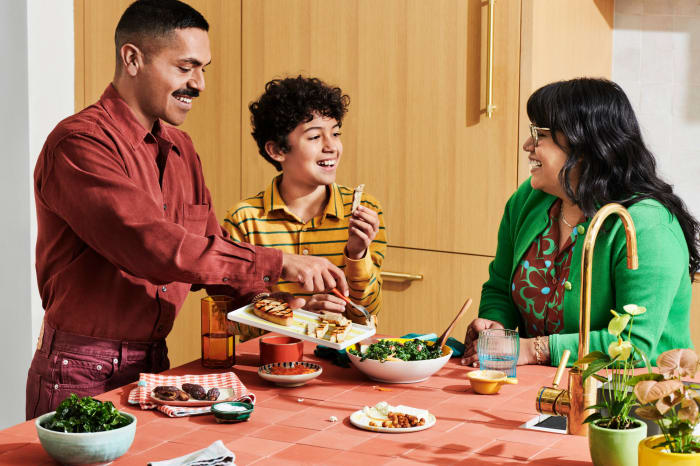 A family of three, including a man, a child, and a woman, are smiling and sitting around a table with food and plants in an indoor kitchen setting.