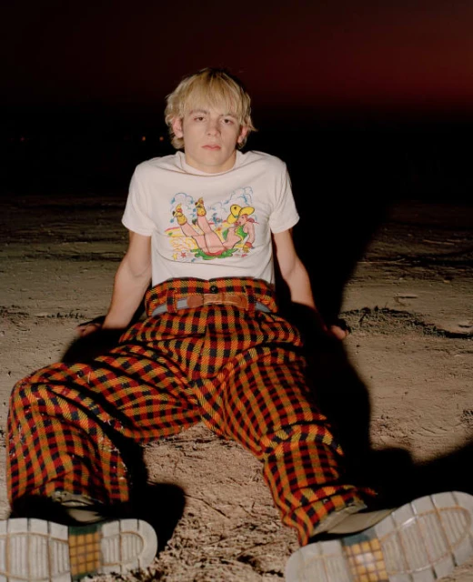 Young male adult sits outdoors in oversized plaid pants and t-shirt with graphic design. Brown sand, dark background, eye-level portrait.