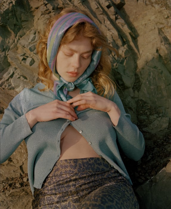 A fair-skinned woman with a headscarf poses outside against a rocky background wearing a blue cardigan and patterned pants.