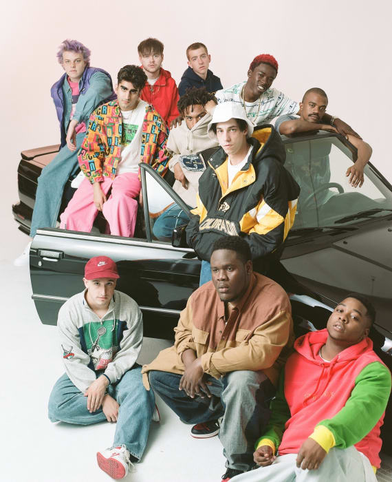 A group of young men casually pose near a black car in a bright photo studio, showcasing diverse streetwear and casual attire.