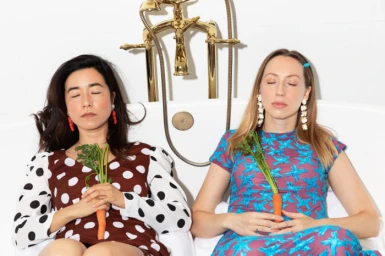 Two women with closed eyes holding whole carrots in a white bathtub styled by Ashley Guerzon