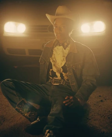 Person in cowboy hat, denim jacket, and jeans seated on the ground in front of a car with glowing headlights.