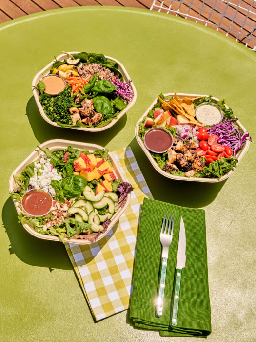 Overhead shot of three bowls of salads with various toppings on a green table, accompanied by silverware on a green napkin.