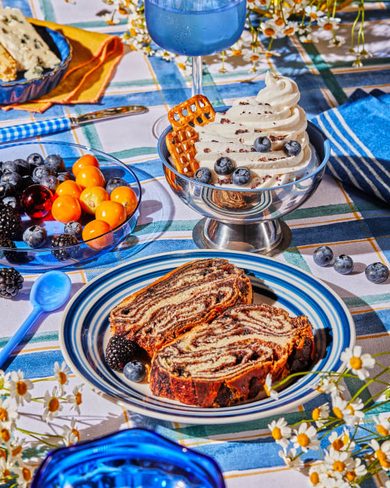 Overhead shot of sponge cake slices, soft-serve ice cream, berries, and a blue drink on a blue and white tablecloth with daisies.