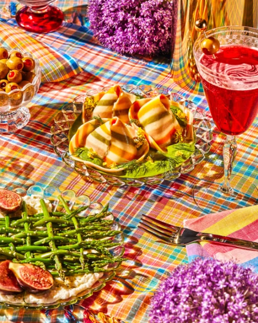 Still life photograph showcasing appetizers, a cocktail, and decorative purple allium flowers arranged on a colourful checkered tablecloth.