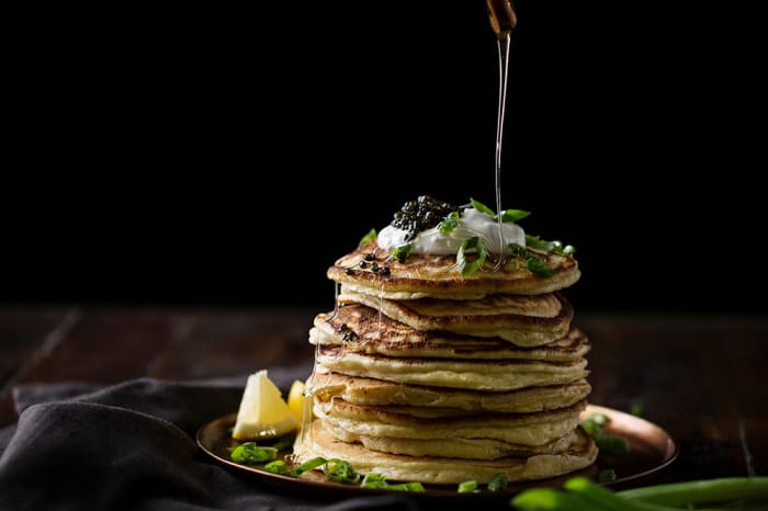 Stack of pancakes topped with caviar and cream, drizzled with honey, set on a dark wood table against a black background.