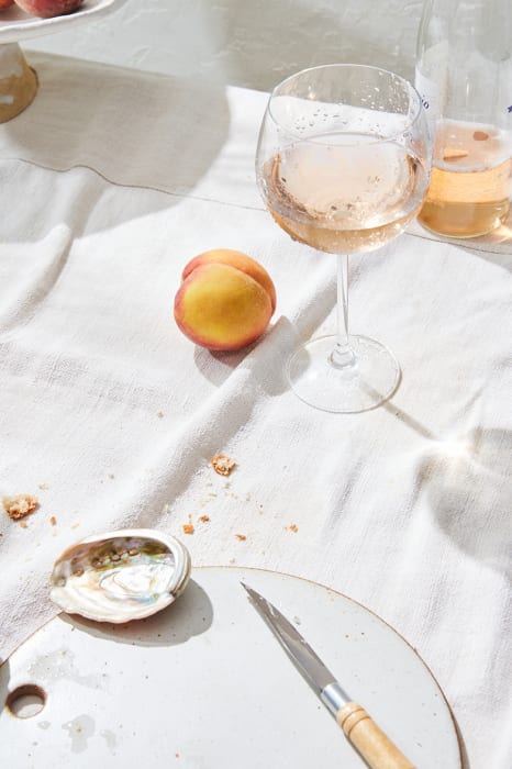 Still life of a wine glass with rosé wine, a peach, an abalone shell, a knife, and a plate on a white linen tablecloth in bright sunlight.