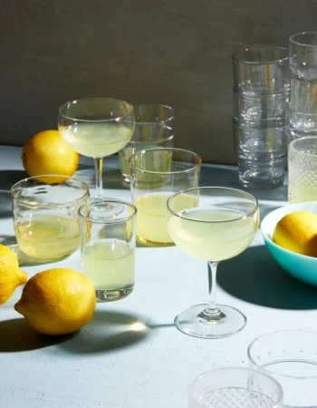 Several glasses of light yellow cocktails are displayed with fresh lemons on a light blue surface with a grey background.
