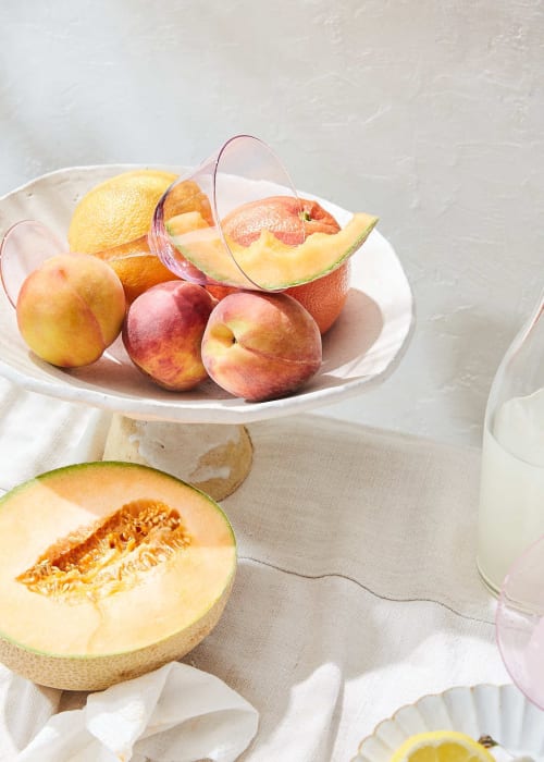 Still life food photography features a cake stand holding peaches, oranges, a cantaloupe, and a glass on a white tablecloth.