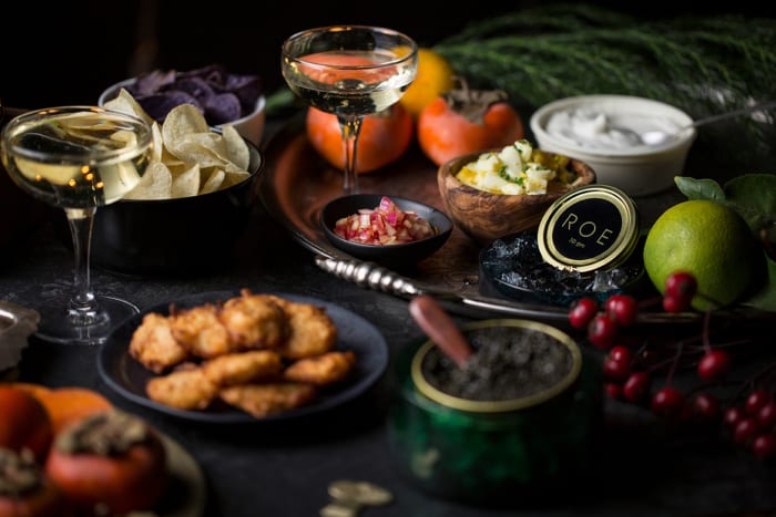 Gourmet food still life: caviar, persimmons, chips, dips, champagne coupes on dark surface with soft focus. Upscale snack arrangement.